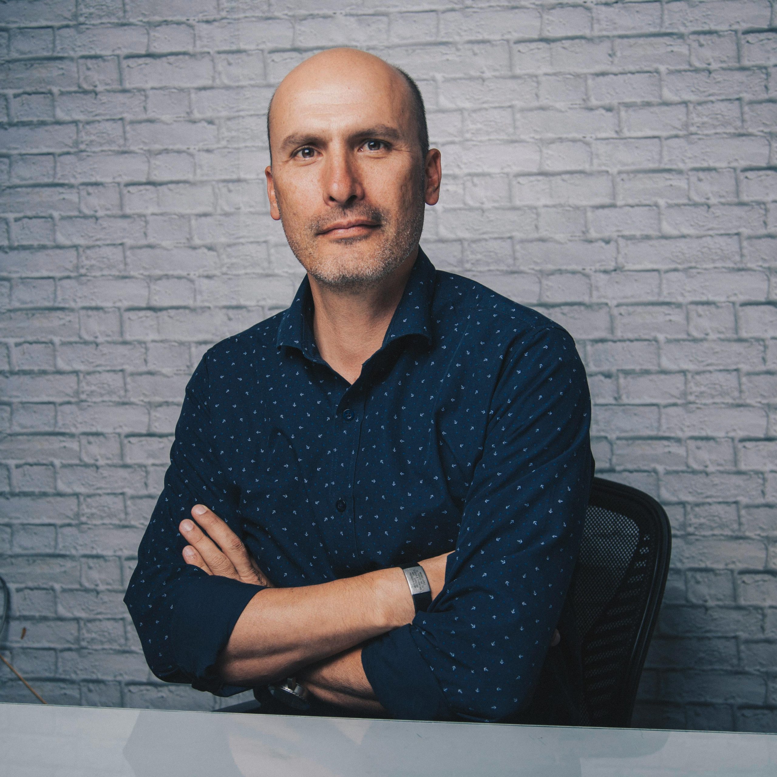 Positive bald male in blue shirt sitting at shiny desk with crossed arms while looking at camera in white room